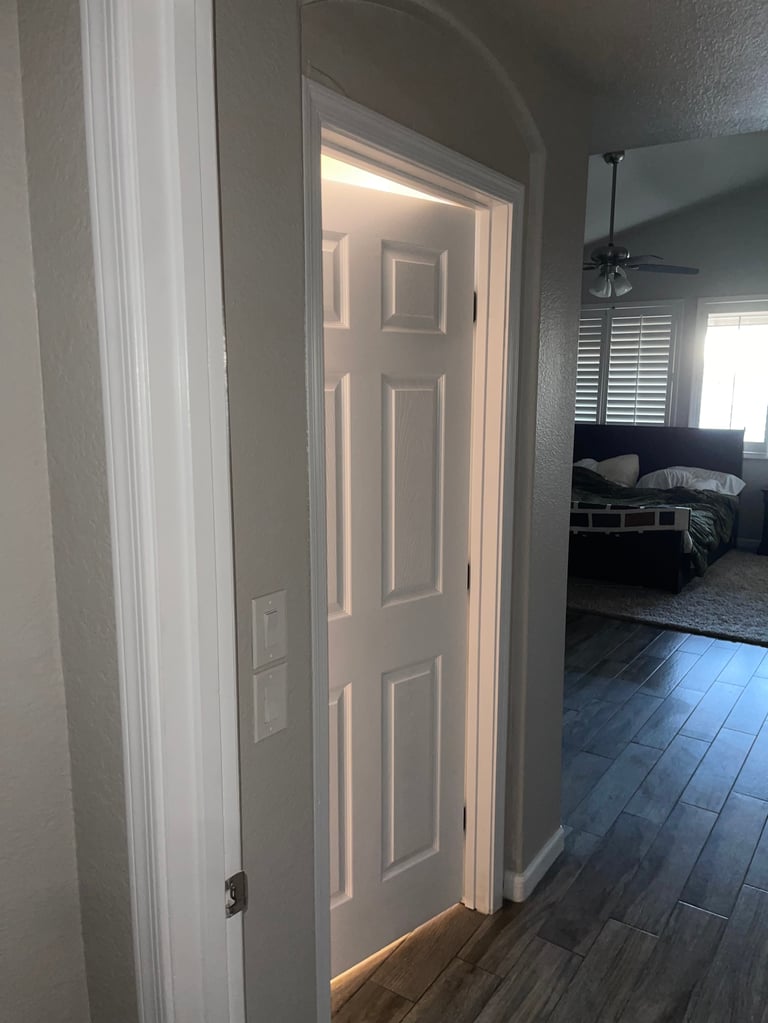 Open white six-panel bedroom door with light glowing from inside room, dark hardwood flooring visible in hallway