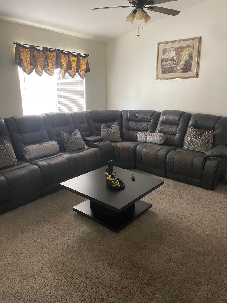 Modern living room with black leather reclining sectional sofa, decorative pillows, black coffee table, window with gold valance, ceiling fan, and framed wall art