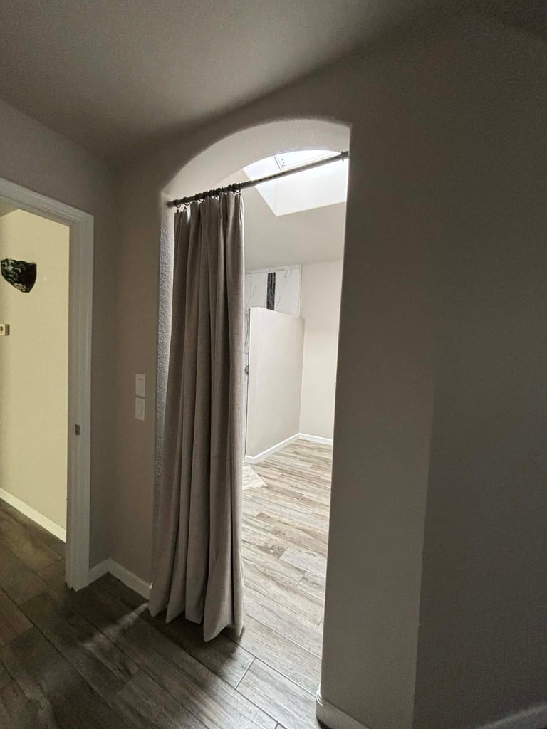 Doorway with beige curtain leading to bright hallway with wood plank flooring and arched ceiling opening