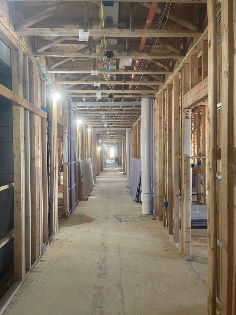 Long hallway under construction with wooden framing, drywall sheets, and exposed ceiling beams lit by work lights